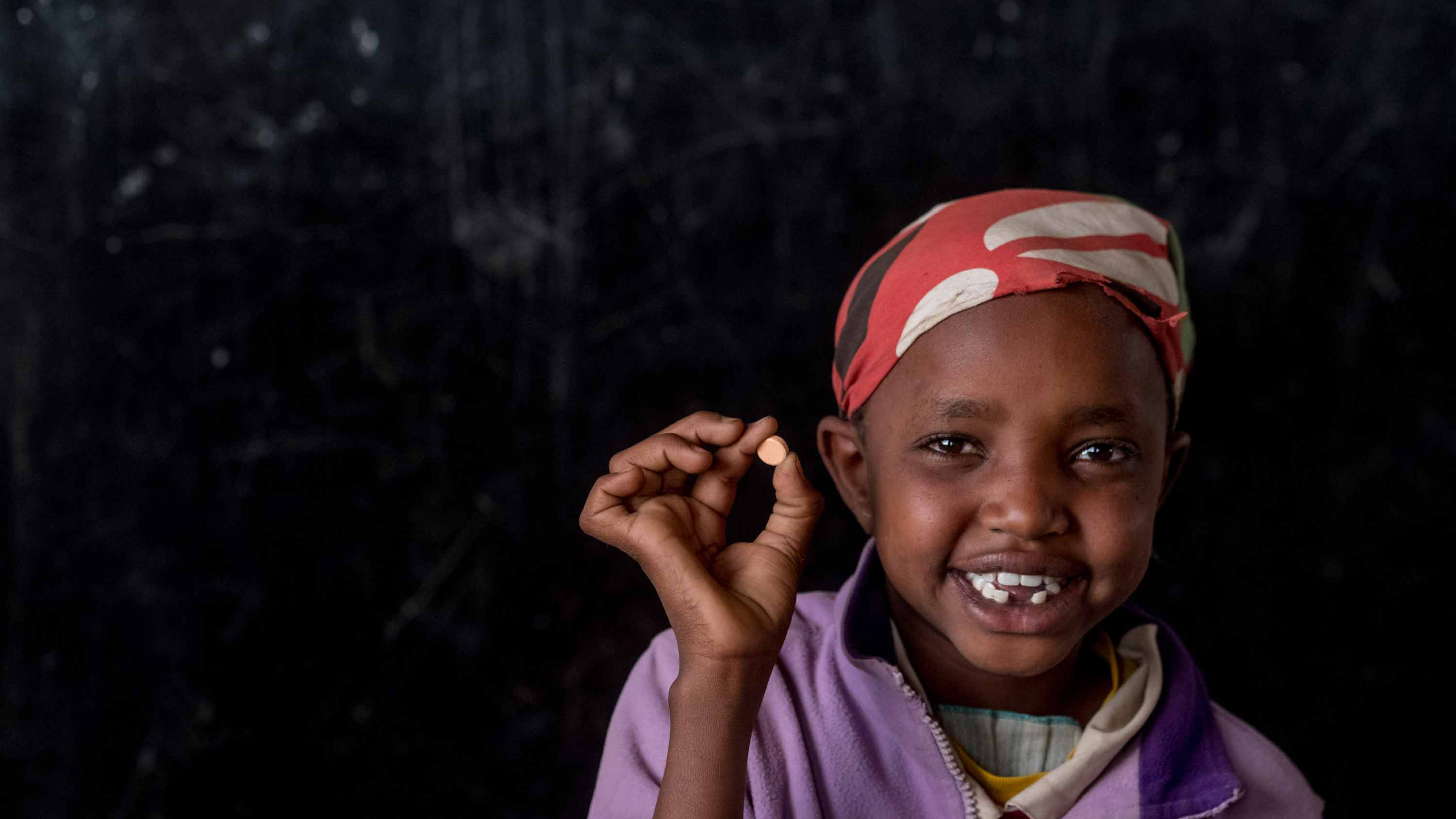 A girl in a red scarf holds up a small white pill.