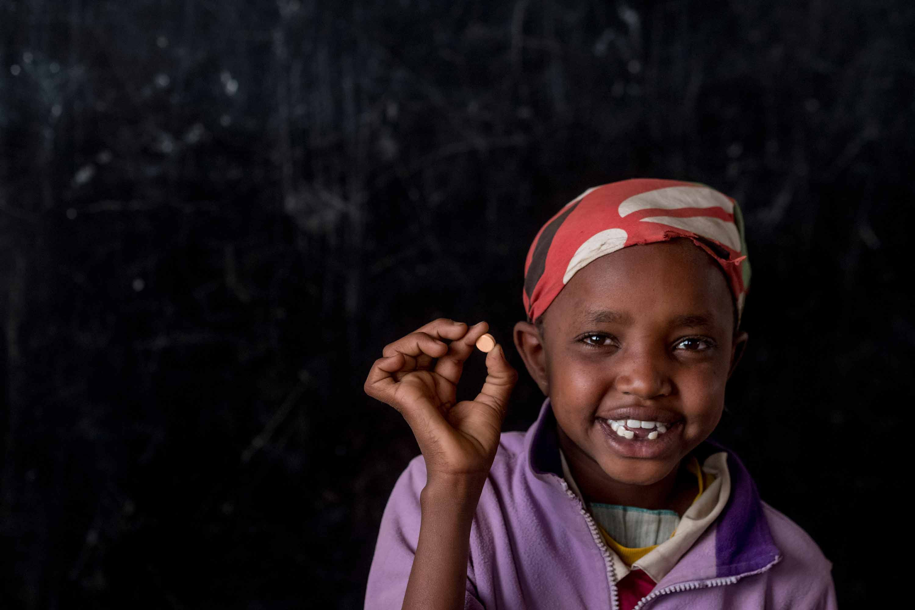 A girl in a red scarf holds up a small white pill.